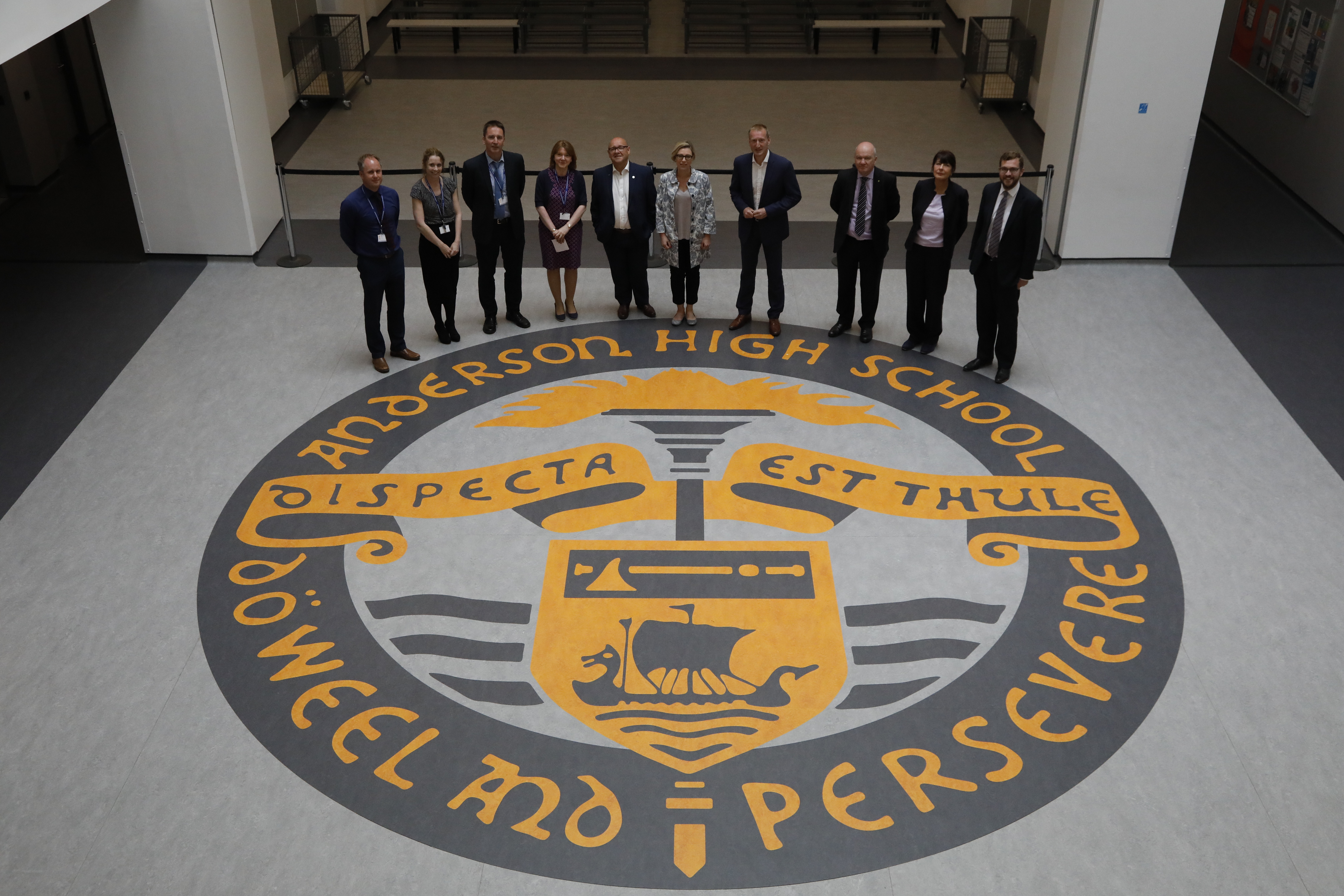 Image is taken from above. It looks down from the first floor to Members of the Education and Skills Committee who are on the ground floor in a large entrance area. Members of the Committee are standing behind a large circular school crest which is embedded in the flooring. The crest has the words 'Anderson High School' and the motto 'Do weel and persevere'.